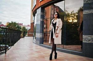 svart hår sexig kvinna i glasögon och kappa poserade mot byggnad med moderna fönster. foto