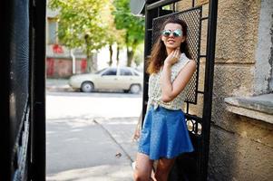 lockigt snygg tjej slitage på blå jeans kjol, blus och solglasögon. porträtt på gatorna i staden. foto