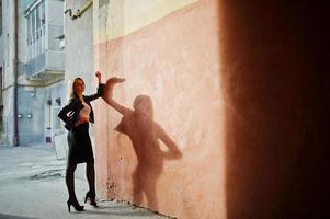 elegant blond flicka bär på svart skinnjacka poserar på gatorna i staden bakgrund gamla väggen med skuggor. foto