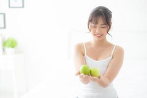 vackert porträtt ung asiatisk kvinna som håller och äter grönt äpple frukt i sovrummet hemma, livsstil för näring flicka hälsosam och omsorg viktminskning, hälsa och välbefinnande koncept. foto