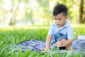 ung liten asiatisk pojke som läser och skriver bok i parken, asia kid läxor och studera på sommaren, barn koppla av med att rita på anteckningsboken i semester, utbildning och utveckling koncept. foto