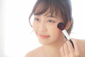 skönhetsporträtt ung asiatisk kvinna leende med ansiktet tittar spegeln applicera makeup med borste kinden i sovrummet, vacker flicka håller rouge, hudvård och kosmetiska koncept. foto