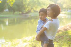 vacker ung asiatisk mamma som bär liten pojke i parken, asien kvinna glad att ha son och kramunge, mamma älskar och omfamnar barn tillsammans på sommaren, morsdagen och familjekonceptet. foto