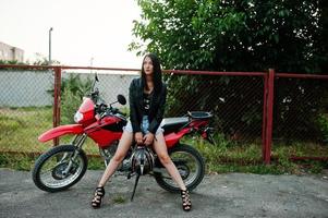 porträtt av en cool och fantastisk kvinna i klänning och svart skinnjacka som sitter på en cool röd motorcykel. foto