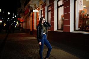 nattporträtt av flickmodell slitage på glasögon, jeans och skinnjacka, med ryggsäck, mot stadens gator. foto