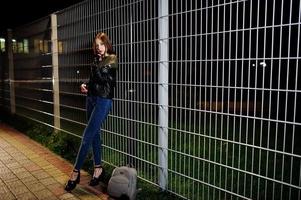 nattporträtt av flicka modell slitage på jeans och skinnjacka mot järn staket. foto