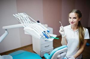 snygg kvinnlig tandläkare poserar med några dentala instrument i handen i vit rock i ett modernt välutrustat skåp. foto