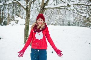 blond flicka i röd halsduk, hatt och jultomtens tröja poserar i parken på vinterdagen. foto