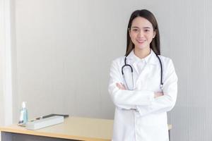 asiatisk kvinna läkare står med armarna i kors glad och leende på sjukhus. iklädd vit mantel och stetoskop. nytt normal- och hälsokoncept. foto