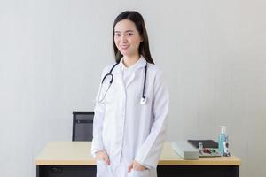en asiatisk kvinnlig läkare med en vit labbrock som står och bär en vit mantel och stetoskop. foto
