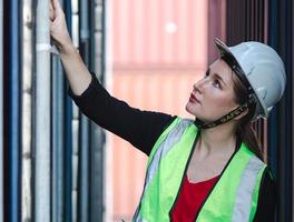 kvinnlig förman arbetare inspekterar containerlåda på kommersiell transport varv. vacker industriingenjör kvinna kontrollerar, undersöker lastcontainer på importexportdistributionslager. foto