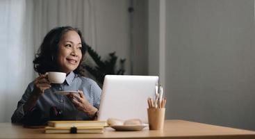 porträtt leende mogen kvinna som njuter av fritid på helgen hemma, använder laptop, glad medelålders kvinna som håller en kopp te eller kaffe, sitter på soffan i vardagsrummet foto