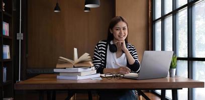 vacker ung asiatisk kvinnlig collegestudent i biblioteket eller kaféet som gör sina läxor och använder en bärbar dator. foto