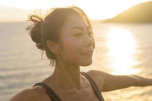 ung asiatisk kvinna i sportkläder gör yoga på klippan vid havet under solnedgången, hälsa och meditation koncept foto