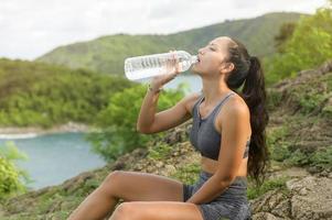 vacker passform kvinna i sportkläder dricksvatten efter att ha tränat på bergstoppen vid havet, hälsa och resekoncept. foto