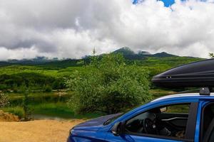 en blå bil med svart takräcke står mot bakgrund av en fjällsjö med berg och moln. foto