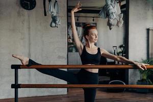 ung atletisk rödhårig ballerina sträcker benet på balett barre i fitnessstudio foto