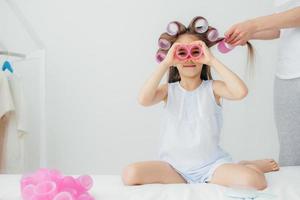 horisontell bild av vacker liten unge har papiljotter på huvudet, leker med dem, kommer att ha underbar frisyr på karneval, isolerad över vit bakgrund. rolig dotter leker tillsammans med mamma foto