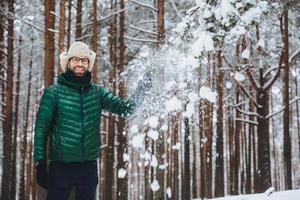 leende förtjusande man klädd i varma kläder, står i vinterskogen, kastar snö i luften, har roligt ensam, har gott humör, uttrycker positiva känslor och känslor. positivitet koncept foto