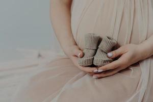 moderskap och graviditet koncept. oigenkännlig gravid kvinna håller små stövlar över magen bär festklänning. ansiktslös framtida mamma foto