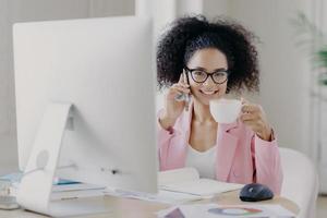 foto av attraktiv lockig, elegant kvinna dricker aromatiskt kaffe, poserar vid skrivbordet med en modern dator som behövs för jobbet, ringer en vän under pausen, diskuterar senaste nyheterna, har ett tandigt leende