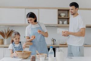 mamma och pappa ger ägg till dottern som lagar deg, är upptagen med att laga mat tillsammans under helgen, har glatt humör, lagar mat. tre familjemedlemmar hemma. föräldraskap och samhörighet koncept foto