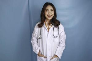 självsäker avslappnad kvinnlig asiatisk läkare som står med händerna i fickorna på sin labbrock och ler tyst mot kameran över blå bakgrund med kopieringsutrymme foto