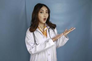 porträtt av en ung asiatisk kvinna läkare, läkare är chock och pekar på ett kopia utrymme isolerat över blå bakgrund foto