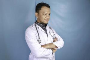 porträtt av en attraktiv självsäker manlig läkare, vänligt leende armar i kors bär vit labbrock stetoskop isolerad blå färg bakgrund foto