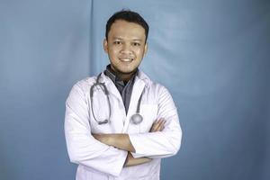 porträtt av en attraktiv självsäker manlig läkare, vänligt leende armar i kors bär vit labbrock stetoskop isolerad blå färg bakgrund foto