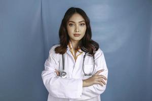 porträtt av en attraktiv självsäker kvinnlig läkare, vänligt leende armar i kors bär vit labbrock stetoskop isolerad blå färgbakgrund foto