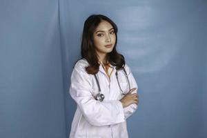 porträtt av en attraktiv självsäker kvinnlig läkare, vänligt leende armar i kors bär vit labbrock stetoskop isolerad blå färgbakgrund foto