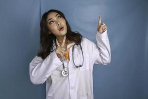 porträtt av en ung asiatisk kvinna läkare, en läkare ler och pekar uppåt på ett kopia utrymme isolerat över blå bakgrund foto