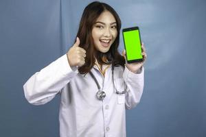 ung asiatisk kvinna läkare pekar och visar grön skärm eller kopiera utrymme på sin smartphone. visar tummen upp eller ok-tecken. foto