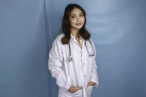 självsäker avslappnad kvinnlig asiatisk läkare som står med händerna i fickorna på sin labbrock och ler tyst mot kameran över blå bakgrund med kopieringsutrymme foto
