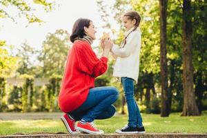 ung mamma i varm stickad röd tröja leker med sin lilla dotter i parken, ger henne löv, njut av soligt höstväder. tillgiven mamma och lilla barn spenderar tid tillsammans utomhus foto