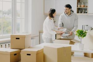 foto av upptaget familjepar packar upp personliga saker från kartonger, klädda i vardagskläder, håller vita tallrikar, poserar i ett rymligt kök med moderna möbler, omgiven av hög med paket