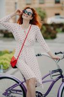 aktiv glad kvinna cyklar i staden, bär snygga solglasögon, har ett charmigt leende, klädd i sommarklänning, njuter av soliga dagar, tillbringar ledig tid aktivt. människor, transporter och livsstil foto