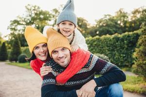 utomhus porträtt av stilig man ge piggyback till sin fru och dotter, bära varma kläder, ha glada uttryck, stödja varandra. familj omfamning utomhus, le glatt in i kameran foto