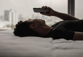 asiatisk man känner sig ledsen på sängen och tittar och fokuserar på smartphone. Internetberoende för sociala medier och koncept för mental hälsa. foto