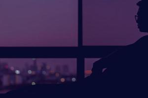 en man som sitter på sin säng ensam på natten och tittar ut på färgglada stadsbokeh-ljus. foto