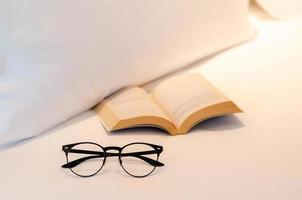 glasögon sätter nära bok på vitt lakan på sängen. avkopplande hemma koncept. foto
