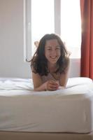 glad ung flicka med mörkt lockigt hår sitter i sängen på morgonen foto