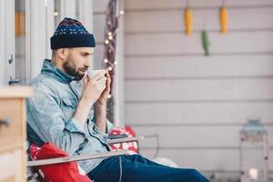 foto av stilig man minns något i livet medan han dricker te eller kaffe, har en tankeväckande blick nedåt, återskapar på terrassen, bär en moderiktig hatt och jeansjacka. fritid och människor koncept