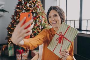 ung italiensk kvinna online bloggare gör selfie med julklapp på bakgrund av julgran foto