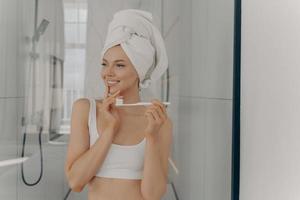 munhygien koncept. frisk vacker kvinna med vit badhandduk på huvudet borstar tänderna under morgonrutinen foto