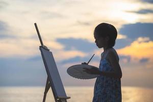 siluett av unge flicka målning på duken på stranden. lyckligt barn flicka rita en bild utomhus foto