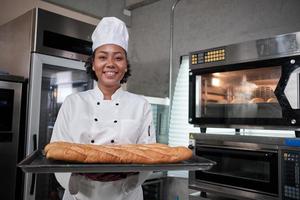 porträtt av afrikansk amerikansk kvinnlig kock i vit matlagningsuniform tittar på kameran med ett glatt leende och stolt med en bricka med baguette i köket, professionella bakverk, färskt bageriyrke. foto