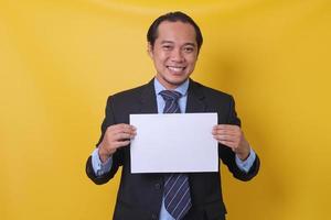 asiatisk ung affärsman i kostym med tomt pappersark isolerad på gul bakgrund med kopia utrymme. håna upp i affärsmannens hand. område för reklam. foto
