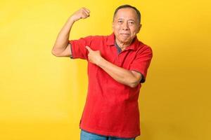 asiatisk pensionär gammal man höja armen direkt finger visa biceps bära röd t-shirt isolerad gul färg bakgrund foto